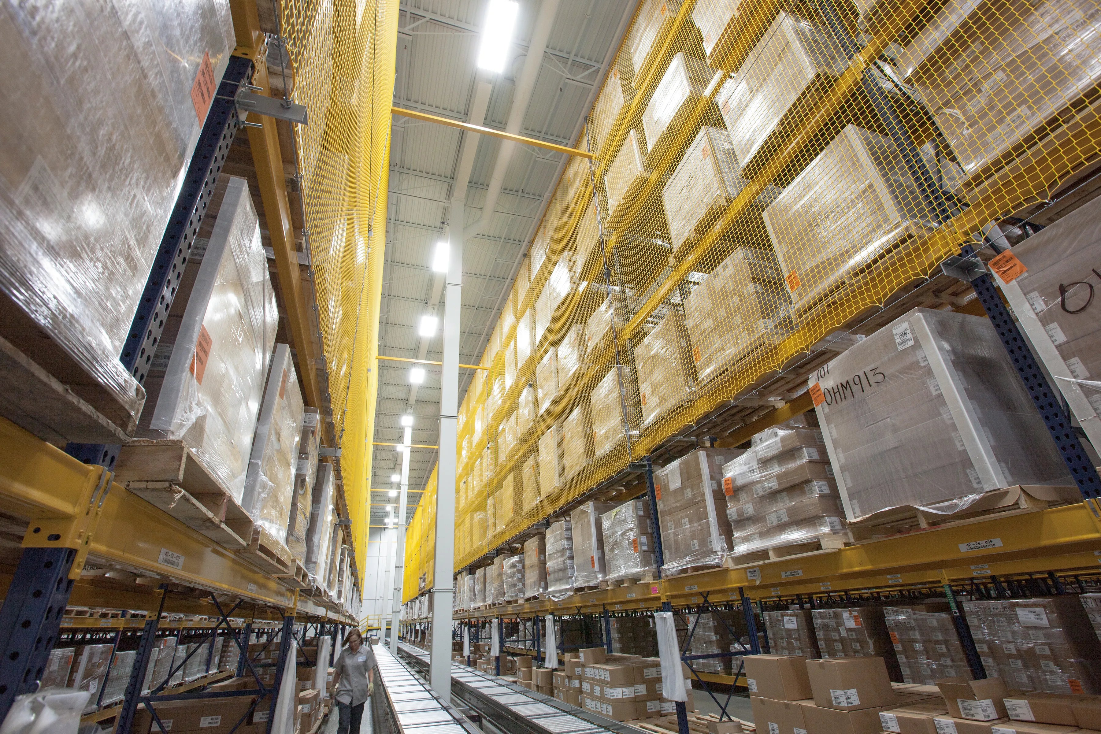 A high-angle view down a narrow aisle in a large warehouse, showing tall yellow and blue shelving stocked with cardboard boxes and palletized goods, protected by mesh nets.